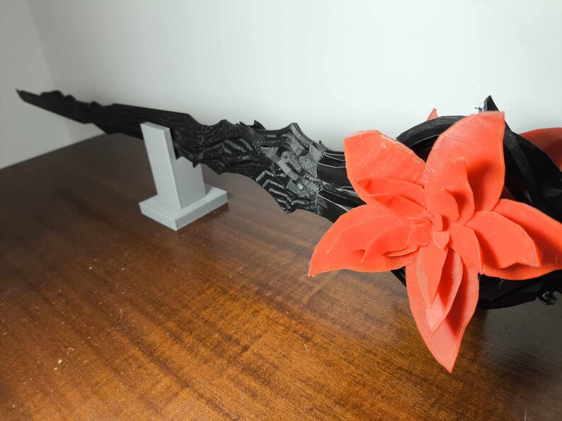 May include: A black, textured, blade-like object is displayed on a gray stand, with a large, orange flower on the right. The objects are on a brown wooden surface. The blade has a jagged edge.
