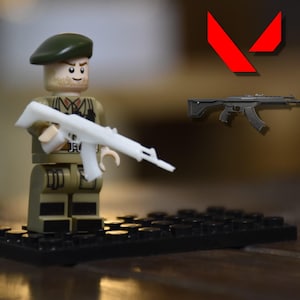 May include: A Lego figurine of a soldier in olive green uniform and a green beret, holding a white rifle. The figurine stands on a black base. A black and gray rifle and a red logo are in the background.