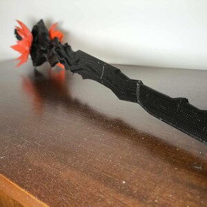 May include: A black, textured wand with a jagged design. The wand's tip features a cluster of orange and black decorative elements. The wand is lying on a dark brown wooden surface.