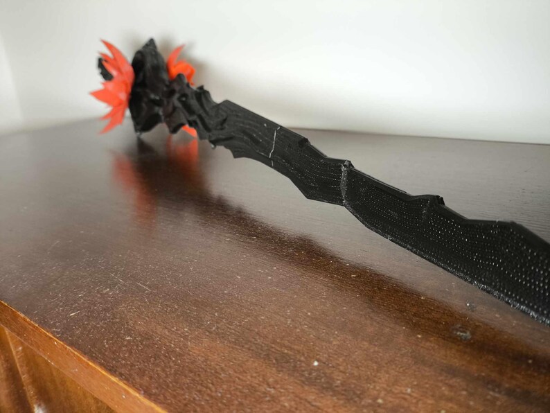 May include: A black, textured wand with a jagged design. The wand's tip features a cluster of orange and black decorative elements. The wand is lying on a dark brown wooden surface.