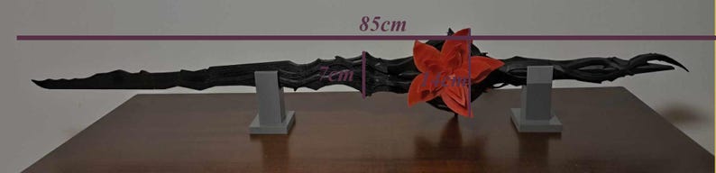May include: A black, ornate staff, 85cm long, with a red flower accent. The staff is displayed on two gray stands. The image shows the staff's intricate design and the flower's vibrant color.