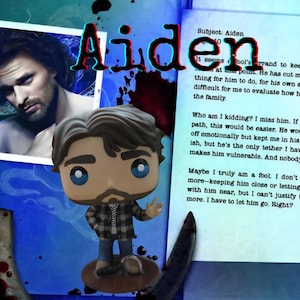 May include: A stylized vinyl figure of a man with brown hair, a beard, and blue eyes, wearing a gray and black plaid shirt and blue pants. The figure is positioned in front of a document titled Subject: Aiden with text and a photograph of a man.