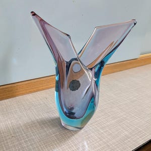 Puede incluir: Un jarrón decorativo de vidrio con una forma y combinación de colores únicas. El jarrón presenta una combinación de vidrio azul, morado y transparente, con una parte superior acampanada. Un artículo de decoración del hogar elegante.