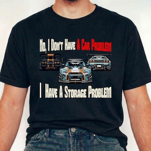Puede incluir: Camiseta negra con un gráfico de tres coches de carreras estilizados. El texto de la camiseta dice: "No, no tengo un problema con el coche, tengo un problema de almacenamiento". Los coches son en tonos de negro, azul, naranja y gris.