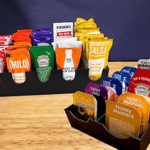 Puede incluir: Organizadores de condimentos negros llenos de varios paquetes de salsa, incluyendo ketchup, mostaza y salsa. Los paquetes están dispuestos en una exhibición escalonada, mostrando una variedad de marcas y sabores. Sobre una superficie de madera.