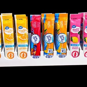 May include: A white plastic organizer holds several packets of drink mix in various flavors. The packets are orange, red, and pink, with text indicating flavors like Orange, Fruit Punch, Citrus, and Raspberry Lemonade. The packets are labeled "Sugar Free."