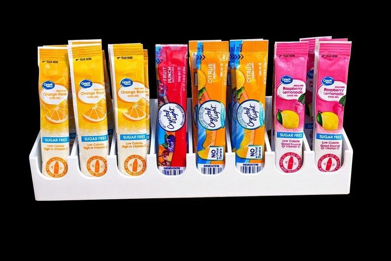 May include: A white plastic organizer holds several packets of drink mix in various flavors. The packets are orange, red, and pink, with text indicating flavors like Orange, Fruit Punch, Citrus, and Raspberry Lemonade. The packets are labeled "Sugar Free."