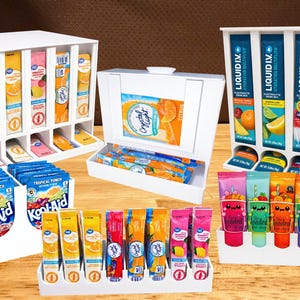 May include: Assortment of drink mix packets and tubes in white display cases. Brands include Crystal Light, Kool-Aid, and Liquid I.V. Flavors include citrus, tropical punch, and various fruit blends. The Loaded Tea Shop tubes are also visible.