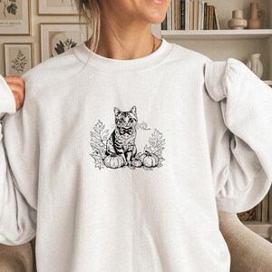 Peut inclure: Un sweat-shirt blanc à col rond avec une illustration au trait noir centrée sur la poitrine. Le motif représente un chat assis portant un nœud papillon, entouré de feuilles d'automne détaillées et de deux petites citrouilles de chaque côté.