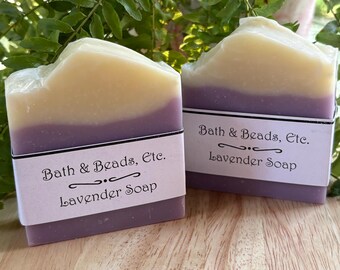 May include: Two rectangular bars of lavender soap featuring a dual-tone design with a creamy white top layer and a solid purple bottom layer. Each bar is wrapped in a white paper label printed with the text Bath & Beads, Etc. Lavender Soap.