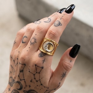 Puede incluir: Anillo dorado con banda rectangular y cara circular con diseño de media luna. El anillo se lleva en una mano con esmalte de uñas negro y tatuajes celestiales, incluyendo constelaciones, planetas y lunas.