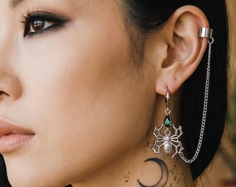 Può includere: Set di polsino e orecchini argentati. L'orecchino presenta un design a ragno con una gemma verde e una luna crescente. Una delicata catena collega il polsino all'orecchino. La persona ha un tatuaggio sul collo di stelle, pianeti e costellazioni.