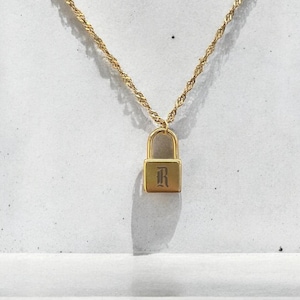 Puede incluir: Un collar de cadena dorada con un pequeño colgante de candado dorado grabado con la letra "R".