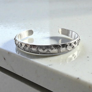 May include: A silver cuff bracelet with an open design. The bracelet features an engraved pattern of crescent moons, stars, and other celestial shapes. The bracelet is approximately 2.5 inches in diameter and is displayed on a white surface.