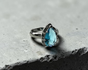 Può includere: Un anello color argento con una singola gemma sfaccettata a forma di pera di colore azzurro, incastonata in una montatura metallica testurizzata con griffe. La fascia è liscia e lucida, in contrasto con la finitura organica e irregolare.
