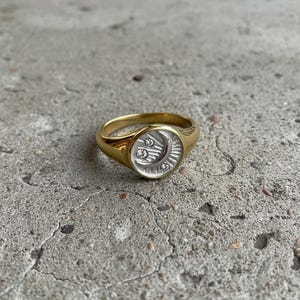 May include: A gold-toned ring featuring a circular face with a silver-toned inlay. The inlay displays a crescent moon and star motif with three small, clear, round-cut stones embedded within the design, set against a textured background.