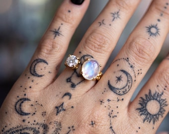 Può includere: Un anello in oro con una pietra chiara rotonda a taglio brillante e una pietra di luna ovale iridescente posizionate fianco a fianco. L'anello presenta una fascia in oro lucido e montature a griffe che fissano saldamente entrambe le pietre.