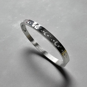 May include: A silver bangle bracelet featuring a repeating pattern of crescent moons and stars. The bracelet has a simple, elegant design. The bracelet is made of a shiny, metallic material. The bracelet's circumference is approximately 18 cm.
