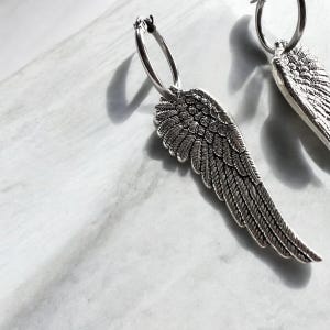 May include: A pair of silver hoop earrings with angel wing charms. The wings are detailed with a textured feather design.