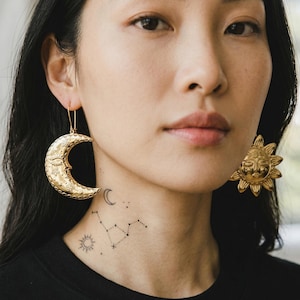 Puede incluir: Pendientes celestiales dorados. Un pendiente es una luna creciente con una cara, el otro es un sol con una cara. Los pendientes los lleva una persona con una camiseta negra y un tatuaje en el cuello de una luna, un sol y una constelación.