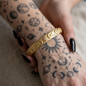 Peut inclure: Bracelet doré orné de motifs célestes : soleils, lunes et étoiles. Le bracelet est orné de petits ornements scintillants. L'image montre également des tatouages de planètes, de lunes et de constellations.