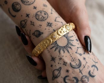 Può includere: Bracciale dorato con disegni celesti, tra cui soli, lune e stelle. Il bracciale è adornato con piccoli abbellimenti scintillanti. L'immagine mostra anche tatuaggi di pianeti, lune e costellazioni sul braccio.