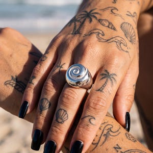 Könnte beinhalten: Ein Ring aus poliertem Silber mit einem markanten, erhabenen Spiraldesign auf der Vorderseite. Der Ring verfügt über eine glatte, abgerundete Ringschiene und eine reflektierende metallische Oberfläche in einer klaren, skulpturalen Form.