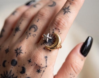 Può includere: Un anello bicolore caratterizzato da una mezzaluna color oro lucido e una stella più piccola color argento. L'anello è indossato su un dito e presenta una finitura metallica liscia su entrambi gli elementi del gioiello.