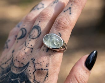 Può includere: Un anello d'argento con una pietra ovale traslucida verde chiaro. L'anello è incastonato in una fascia d'argento lucida. Lo sfondo è sfocato, con l'attenzione sull'anello e sulla mano.