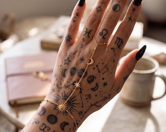 Puede incluir: Una cadena de mano dorada con un diseño de eslabones delicados que incluye un dije de sol en la muñeca, un dije de estrella en el centro y un dije de media luna cerca del lazo para el dedo. La joya está hecha de cadenas metálicas finas.