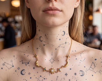 Può includere: Una collana color oro con un ciondolo centrale a forma di sole con una pietra chiara, affiancato da due ciondoli a mezzaluna e piccoli dettagli a stella. La catenina è sottile e metallica, poggiata su una pelle decorata con tatuaggi celesti.