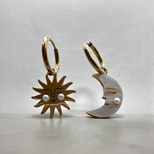 Puede incluir: Pendientes de aro dorados. Un pendiente presenta un diseño de sol con una cara y detalles de perlas. El otro pendiente tiene un diseño de luna con detalles de perlas. Los pendientes están sobre un fondo gris claro.