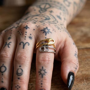 May include: Two safety pin rings, one gold-coloured and one silver-coloured, are stacked on a finger. The rings are set against a background of tattooed skin and a wooden surface. The gold ring is on top of the silver ring.
