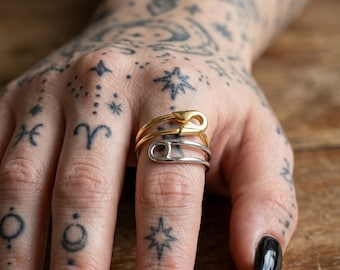 Può includere: Due anelli a forma di spilla da balia, uno dorato e uno argentato, sono impilati su un dito. Gli anelli sono posizionati su uno sfondo di pelle tatuata e una superficie di legno. L'anello dorato è sopra l'anello argentato.