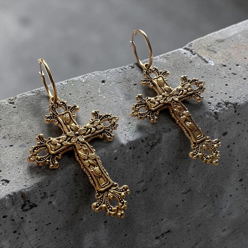 Puede incluir: Un par de pendientes de aro dorados con colgantes de cruz de estilo filigrana. Cada cruz presenta intrincados detalles florales y de volutas en relieve por toda la superficie met&aacute;lica, suspendida de un cierre de aro circular sencillo.