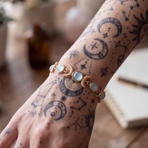 May include: A gold-toned bracelet featuring a series of alternating crescent moon charms and round, pale blue cabochon stones. The bracelet is designed with a linked chain structure, showcasing a celestial motif that connects each moon and stone element.