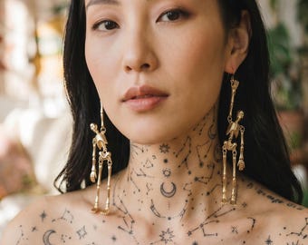 Può includere: Orecchini a scheletro color oro. Gli orecchini sono lunghi e pendenti, con un design a scheletro dettagliato. La persona nell'immagine ha un tatuaggio di costellazione sul collo e sul petto.