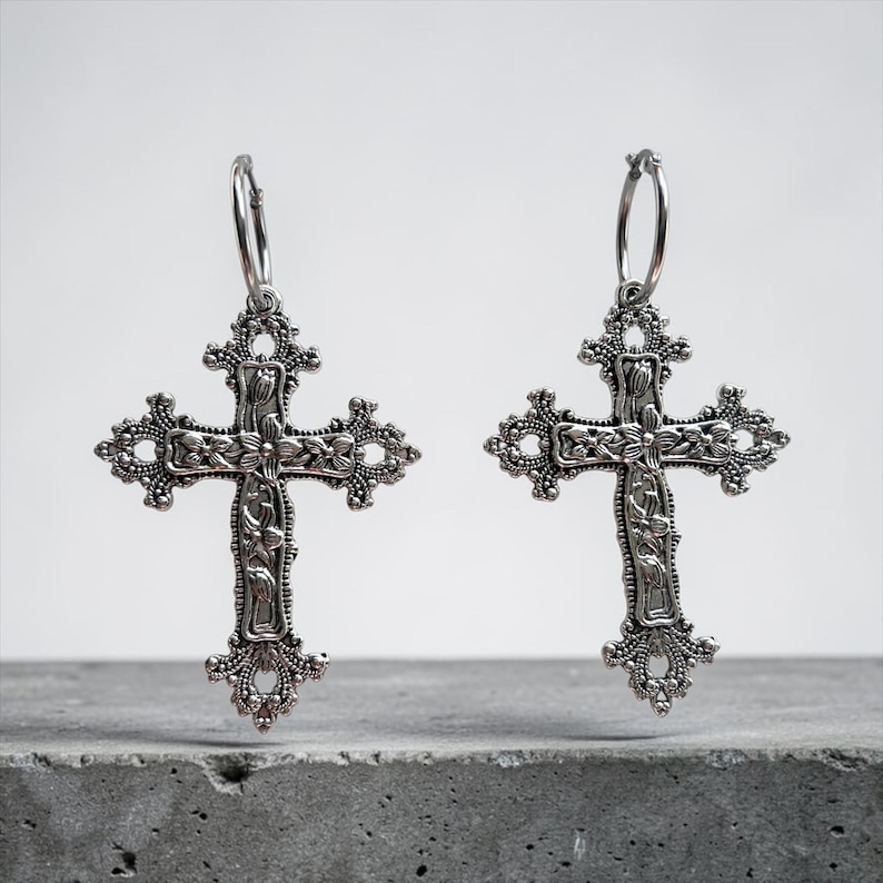 Puede incluir: Un par de pendientes de tono plateado con colgantes de cruz ornamentados suspendidos de aros circulares. Las cruces presentan intrincados grabados florales y bordes de filigrana decorativos, mostrando una textura met&aacute;lica detallada en todo el dise&ntilde;o.