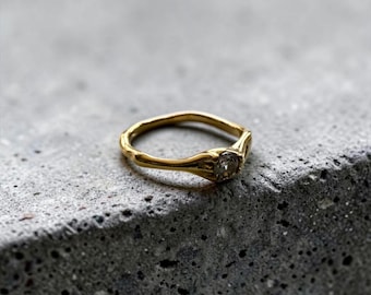Può includere: Un anello in oro con una fascia dalla forma organica e irregolare, caratterizzato da una singola piccola pietra preziosa rotonda e trasparente incastonata a griffe. La superficie metallica lucida e riflettente mette in risalto il design.