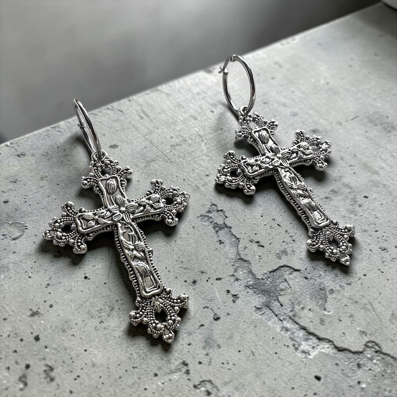 Puede incluir: Un par de pendientes de aro plateados con colgantes en forma de cruz ornamentada. Cada cruz presenta detalles florales y de volutas en relieve por toda su superficie, con elementos decorativos de filigrana en los extremos de los brazos.