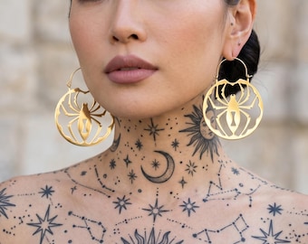 Può includere: Orecchini a cerchio dorati a forma di ragno. La persona indossa un top nero e intricati tatuaggi celesti sul collo e sul petto, tra cui stelle, costellazioni e un sole.