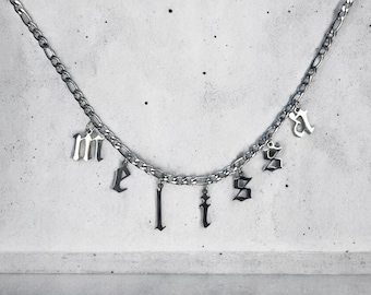 Può includere: Una collana color argento con un design a catena. La collana presenta più ciondoli pendenti che compongono la parola "melts" in caratteri gotici. Lo sfondo è grigio neutro.