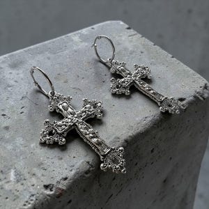 Puede incluir: Un par de pendientes de aro en tono plateado con colgantes de cruz detallados y ornamentados. Cada cruz presenta un intrincado trabajo de metal estilo filigrana con superficies texturizadas y adornos decorativos en los extremos.