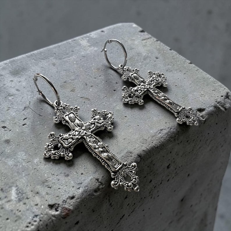 Puede incluir: Un par de pendientes de aro en tono plateado con colgantes de cruz detallados y ornamentados. Cada cruz presenta un intrincado trabajo de metal estilo filigrana con superficies texturizadas y adornos decorativos en los extremos.