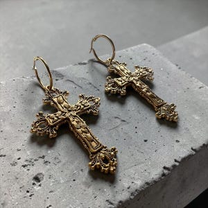 Puede incluir: Un par de pendientes de aro en tono dorado con colgantes de cruz de estilo filigrana ornamentada. Cada cruz presenta detalles florales intrincados y volutas decorativas en los extremos de los brazos, suspendidas de un aro de metal simple.