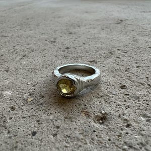 May include: A silver ring with a textured band and a round, yellow gemstone. The ring has a unique, organic design, with the gemstone set in a bezel setting. The ring is displayed on a gray surface.