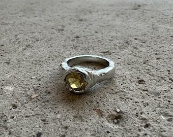 Puede incluir: Un anillo plateado con una banda texturizada y una gema amarilla redonda. El anillo tiene un diseño orgánico único, con la gema engastada en un bisel. El anillo se muestra sobre una superficie gris.