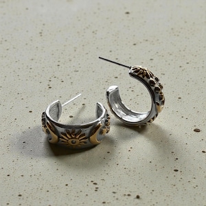 Puede incluir: Un par de pendientes de aro plateados con detalles dorados. Los pendientes presentan un diseño de sol y luna. Los detalles dorados están en los bordes exteriores de los aros. Los pendientes están sobre una superficie gris claro.