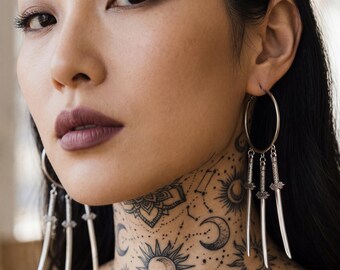 Può includere: Primo piano di una persona che indossa orecchini a cerchio argentati con tre ciondoli a forma di spada. La persona ha un tatuaggio dettagliato di disegni celesti sul collo e sul petto.