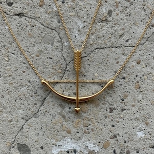 May include: A gold-colored necklace featuring a bow and arrow pendant. The bow is curved, and the arrow is centered, with a small heart at the arrow's tip. The necklace chain is delicate and visible against the gray background.